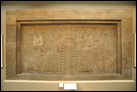 Символическая сцена. Нимруд, ок. 865-860 гг. до Р.Х. Британский музей. WA 124531. Царь Ашшурнацирпал II изображен дважды — в ритуальной одежде (слева) и с царским жезлом (справа). Напротив него находится священное дерево, которое, возможно, символизирует жизнь, а царь совершает поклонение божеству в виде парящего диска с крыльями. Этот бог, который может быть богом солнца Шамашом, в одной руке держит кольцо —  древний месопотамский символ дарованного богом царствования. За царем с обеих сторон находятся духи-хранители. Эта симметричная картина, исполненная символизма, находилась за царским троном. Была также другая, которая находилась напротив главной двери тронного зала. Похожие сцены занимали важные местоположения и в некоторых других ассирийских дворцах; они также вышивались на царских одеждах.