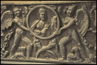 Стенка саркофага. Мрамор. Рим. Конец III в. Эрмитаж. А 1126. В центре — изображение умершей в медальоне, который поддерживают два Эрота. По сторонам — два гения плодородия.